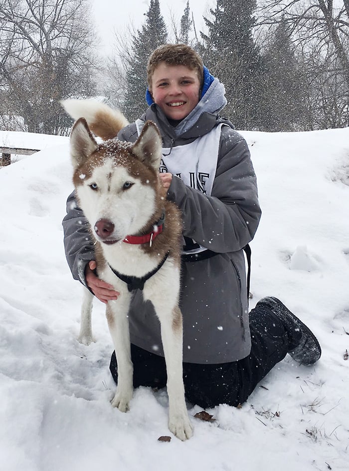 A portrait photo of Dominic, a 13-year-old race dog handler posing with Nanook the husky.