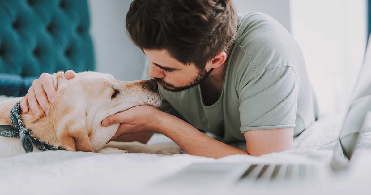A young man lying on a bed kissing his dog on the nose.