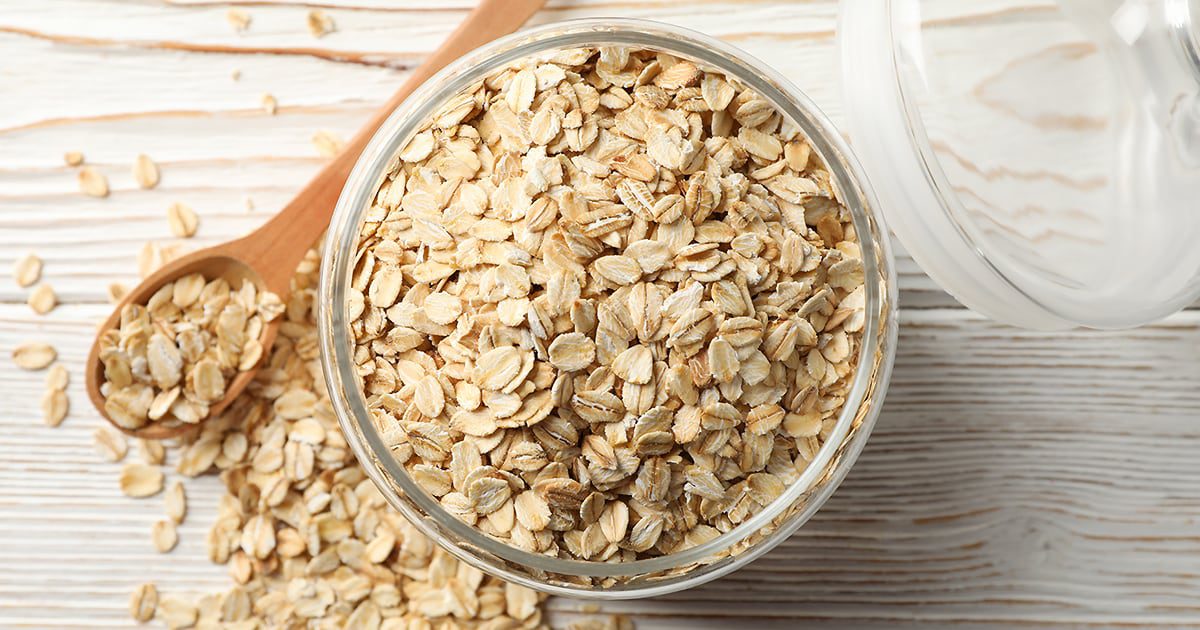 A glass bowl full of oats sitting on a table next to a wooden spoon and a pile of more oats.