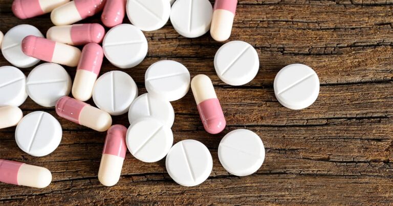 A scattered pile of round white tablets and pink-and-white capsules lying on a wooden table.