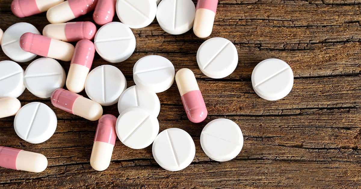 A scattered pile of round white tablets and pink-and-white capsules lying on a wooden table.